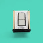 This image depicts a standard white tactile card that is 4 by 6 inch size and displays two white ceramic tiles square centered between a black rectangular outline, labeled on the bottom is the word "Bathroom"