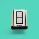 This image depicts a standard white tactile card that is 4 by 6 inch size and displays two white ceramic tiles square centered between a black rectangular outline, labeled on the bottom is the word "Bathroom"