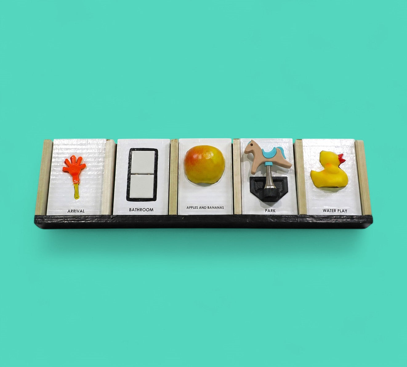 This image depicts five standard 4 by 6 inch tactile cards in a row. From left to right the cards display an orange hand with a yellow handle labeled Arrival, two white ceramic square buttons between a black rectangular outline labeled Bathroom, a yellow and red round apple labeled Apple and Bananas, a figure of a horse attached to a spring labeled Park and a side facing yellow rubber duck with an orange beak labeled Water Play.