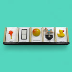This image depicts five standard 4 by 6 inch tactile cards in a row. From left to right the cards display an orange hand with a yellow handle labeled Arrival, two white ceramic square buttons between a black rectangular outline labeled Bathroom, a yellow and red round apple labeled Apple and Bananas, a figure of a horse attached to a spring labeled Park and a side facing yellow rubber duck with an orange beak labeled Water Play.