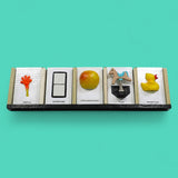 This image depicts five standard 4 by 6 inch tactile cards in a row. From left to right the cards display an orange hand with a yellow handle labeled Arrival, two white ceramic square buttons between a black rectangular outline labeled Bathroom, a yellow and red round apple labeled Apple and Bananas, a figure of a horse attached to a spring labeled Park and a side facing yellow rubber duck with an orange beak labeled Water Play.