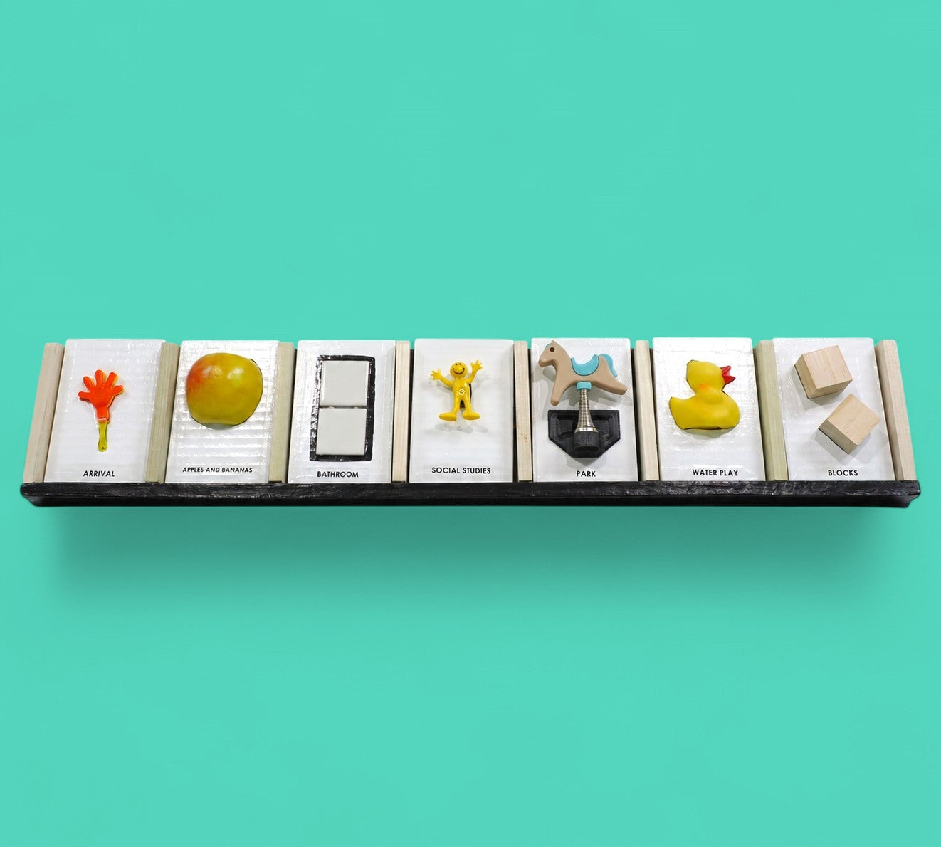 This image displays 7 standard white tactile cards that are 4 by 6 inches in size. From left to right, the cards display an orange hand with a yellow handle labeled Arrival, a yellow apple labeled Apples and Bananas, two white ceramic square buttons centered between a black rectangle labeled Bathroom, a yellow human figure labeled Social Studies, a horse figure attached to a spring labeled Park, a side facing yellow rubber duck labeled Water Play, and two plain square wooden blocks labeled Blocks.
