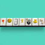 This image displays 7 standard white tactile cards that are 4 by 6 inches in size. From left to right, the cards display an orange hand with a yellow handle labeled Arrival, a yellow apple labeled Apples and Bananas, two white ceramic square buttons centered between a black rectangle labeled Bathroom, a yellow human figure labeled Social Studies, a horse figure attached to a spring labeled Park, a side facing yellow rubber duck labeled Water Play, and two plain square wooden blocks labeled Blocks.