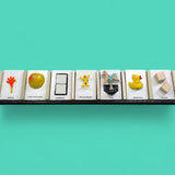 This image displays 7 standard white tactile cards that are 4 by 6 inches in size. From left to right, the cards display an orange hand with a yellow handle labeled Arrival, a yellow apple labeled Apples and Bananas, two white ceramic square buttons centered between a black rectangle labeled Bathroom, a yellow human figure labeled Social Studies, a horse figure attached to a spring labeled Park, a side facing yellow rubber duck labeled Water Play, and two plain square wooden blocks labeled Blocks.