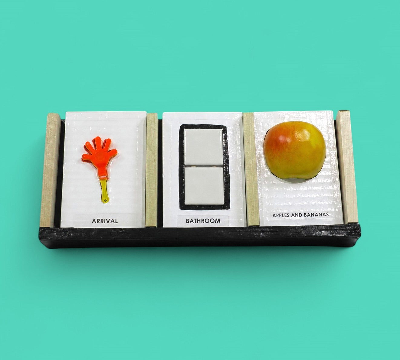 This image depicts 3 standard tactile cards that are 4 by 6 inches in size. The cards from left to right display an orange hand with a yellow handle labeled Arrival, then two square ceramic white buttons centered in a black rectangular outline labeled Bathroom, and a red and yellow apple labeled Apples and Bananas.