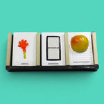 This image depicts 3 standard tactile cards that are 4 by 6 inches in size. The cards from left to right display an orange hand with a yellow handle labeled Arrival, then two square ceramic white buttons centered in a black rectangular outline labeled Bathroom, and a red and yellow apple labeled Apples and Bananas.