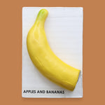 The Apples & Banana Banana symbol has a half of a faux large, yellow banana embedded on a white card. The banana's curved shape draws the viewer's attention, while its vibrant yellow hue adds a pop of color. Labeled "Apples & Banana Banana" in black font at the bottom of symbol on white backing. The background of the symbol is white.