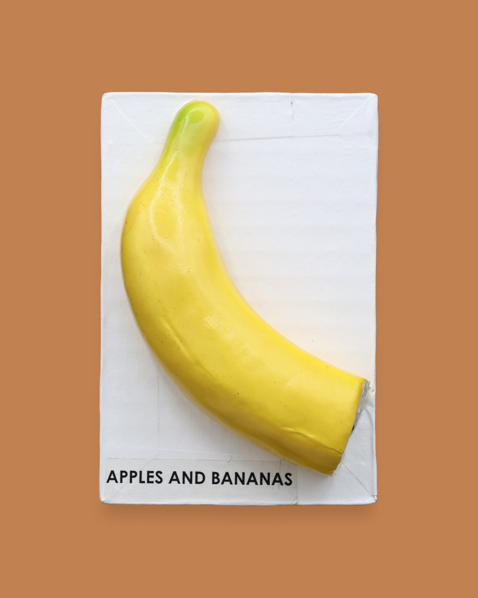 The Apples & Banana Banana symbol has a half of a faux large, yellow banana embedded on a white card. The banana's curved shape draws the viewer's attention, while its vibrant yellow hue adds a pop of color. Labeled "Apples & Banana Banana" in black font at the bottom of symbol on white backing. The background of the symbol is white.