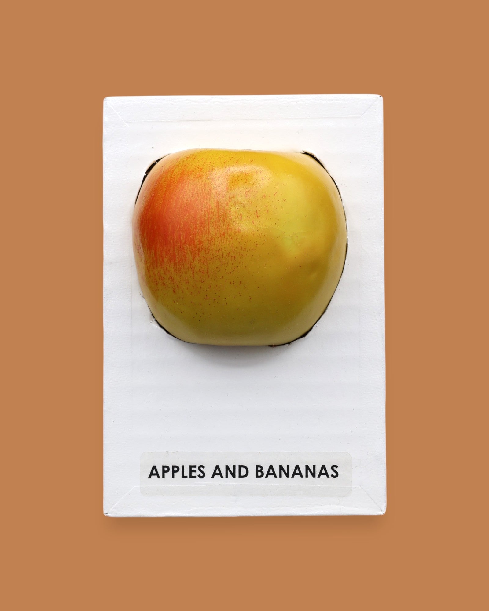 The Apples and Banana symbol with a red faux apple embedded in the center. Labeled "Apples & Banana Apple" in black font at the bottom of symbol on white backing. The background of the symbol is white.