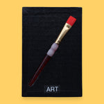 The Art symbol is crafted with a durable plastic paintbrush, the paintbrush handle color may vary. The paintbrush is embedded diagonally. Labeled "Art" in black font at the bottom of symbol on white backing. For black high contrast Art symbol the backing is black with a varied high contrast colored paintbrush. 
