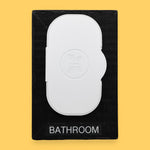 The Bathroom-wipes-lid symbol has a lid of freshwiped embedded vertically in the center of the symbol. Labeled "Bathroom" in black font at the bottom of symbol on white backing. The background of the symbol is white.