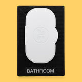 The Bathroom-wipes-lid symbol has a lid of freshwiped embedded vertically in the center of the symbol. Labeled "Bathroom" in black font at the bottom of symbol on white backing. The background of the symbol is white.