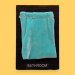 The Bathroom Changing Area symbol has a light blue velvet pouch sewn at the top with a black string embedded just beneath it, leaving the pouch loose to the touch. Labeled "Changing Area" in black font at the bottom of symbol on white backing. The background of the symbol is white.