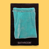 The Bathroom Changing Area symbol has a light blue velvet pouch sewn at the top with a black string embedded just beneath it, leaving the pouch loose to the touch. Labeled "Changing Area" in black font at the bottom of symbol on white backing. The background of the symbol is white.