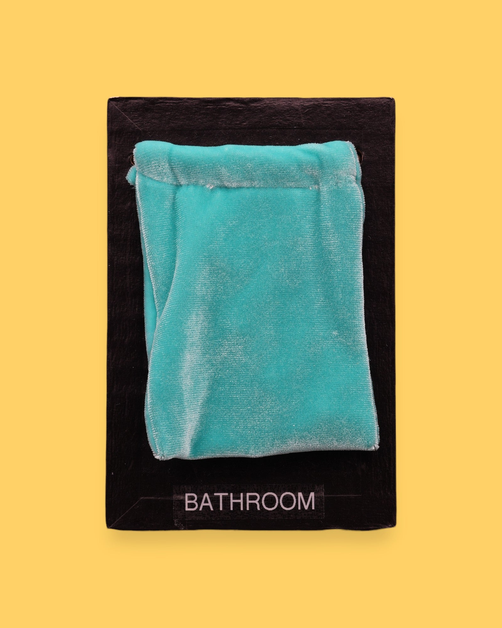 The Bathroom Changing Area symbol has a light blue velvet pouch sewn at the top with a black string embedded just beneath it, leaving the pouch loose to the touch. Labeled "Changing Area" in black font at the bottom of symbol on white backing. The background of the symbol is white.