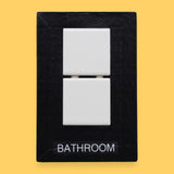 The Bathroom-tile symbol has two square, white, ceramic tiles are embedded vertically in the center of the card. Labeled "Bathroom" in black font on white backing. The background of the symbol is white.