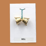 The Bell with two small golden-plated metal bells embedded sided by side with a green interlocked wire holding the bells together. Labeled "Bell" in black font at the bottom of symbol on white backing. The background of the symbol is white.