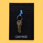 The Car Ride symbol has a golden-plated metal key with a blue rubber handle embedded in the center with white backing. Labeled "Carride" in white font at the bottom of symbol on clear backing. The background of the symbol is black.