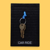 The Car Ride symbol has a golden-plated metal key with a blue rubber handle embedded in the center with white backing. Labeled "Carride" in white font at the bottom of symbol on clear backing. The background of the symbol is black.