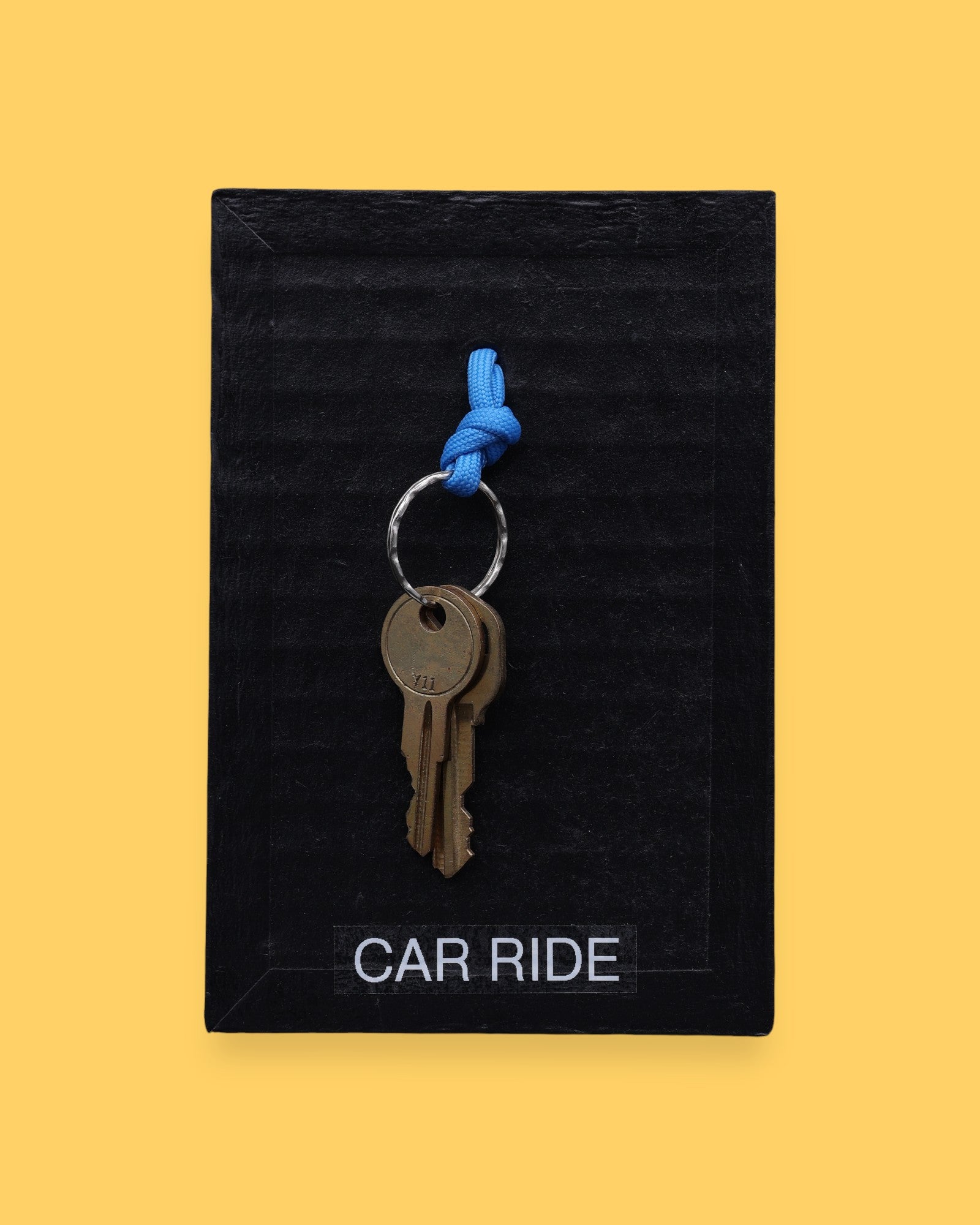 The Car Ride symbol has a golden-plated metal key with a blue rubber handle embedded in the center with white backing. Labeled "Carride" in white font at the bottom of symbol on clear backing. The background of the symbol is black.