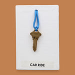 The Car Ride symbol has a golden-plated metal key with a blue rubber handle embedded in the center with white backing. Labeled "Carride" in white font at the bottom of symbol on clear backing. The background of the symbol is black.