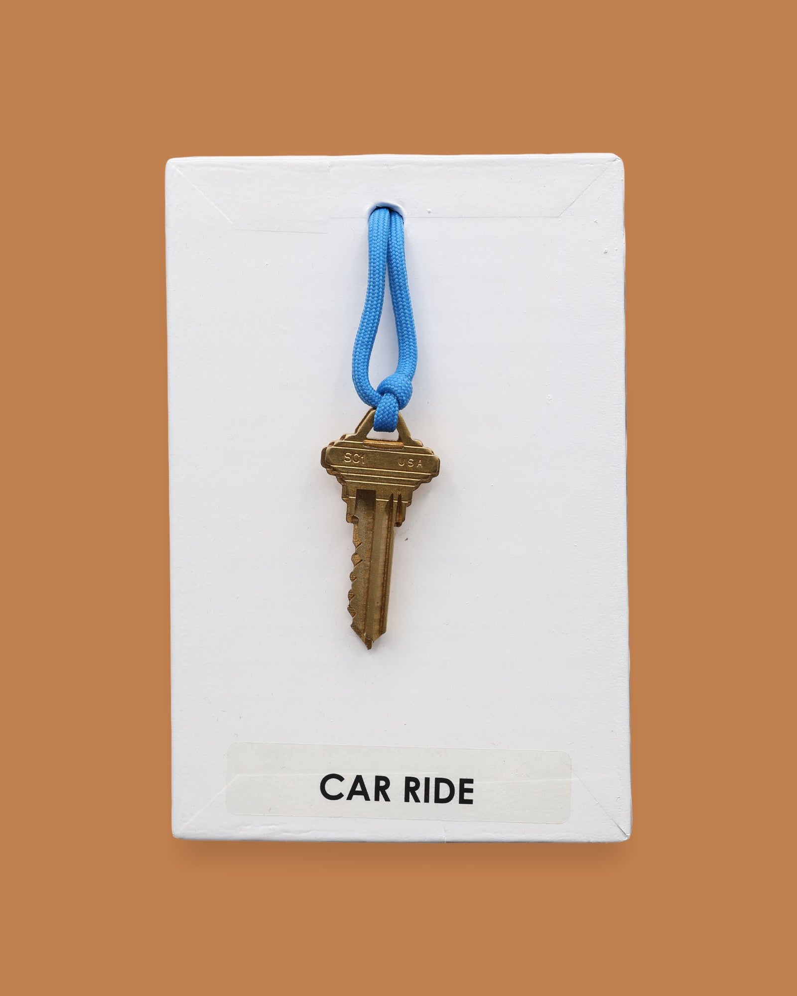 The Car Ride symbol has a golden-plated metal key with a blue rubber handle embedded in the center with white backing. Labeled "Carride" in white font at the bottom of symbol on clear backing. The background of the symbol is black.