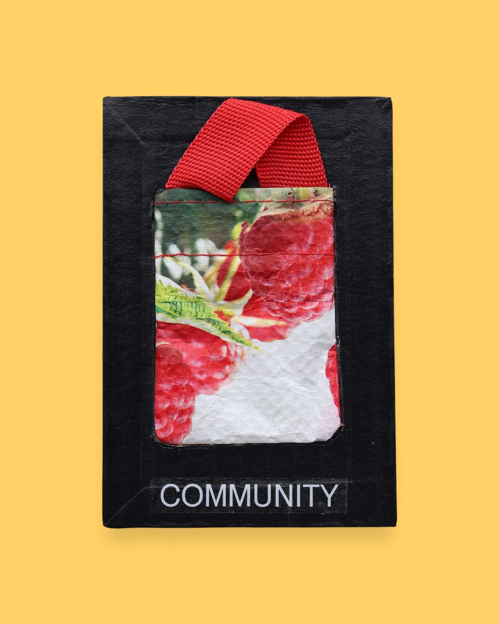 The Community symobl has a small textured bag with red raspberries printed on it, attached by a red strap. Labeled "Community" with white font at the bottom of symbol on clear backing. The background of the symbol is black.