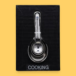 The Cooking symbol has cooking/measuring spoons embedded vertically in the center. Labeled "Cooking" in black font and white backing at the bottom of symbol.. The background of the card is white.