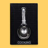 The Cooking symbol has cooking/measuring spoons embedded vertically in the center. Labeled "Cooking" in black font and white backing at the bottom of symbol.. The background of the card is white.