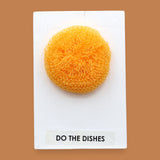 The Do The Dishes symbol with a textured cleaning sponge embedded horizontally. Labeled "Do The Dishes" in black font on white backing at the bottom of the symbol. The background of the symbol is white. 