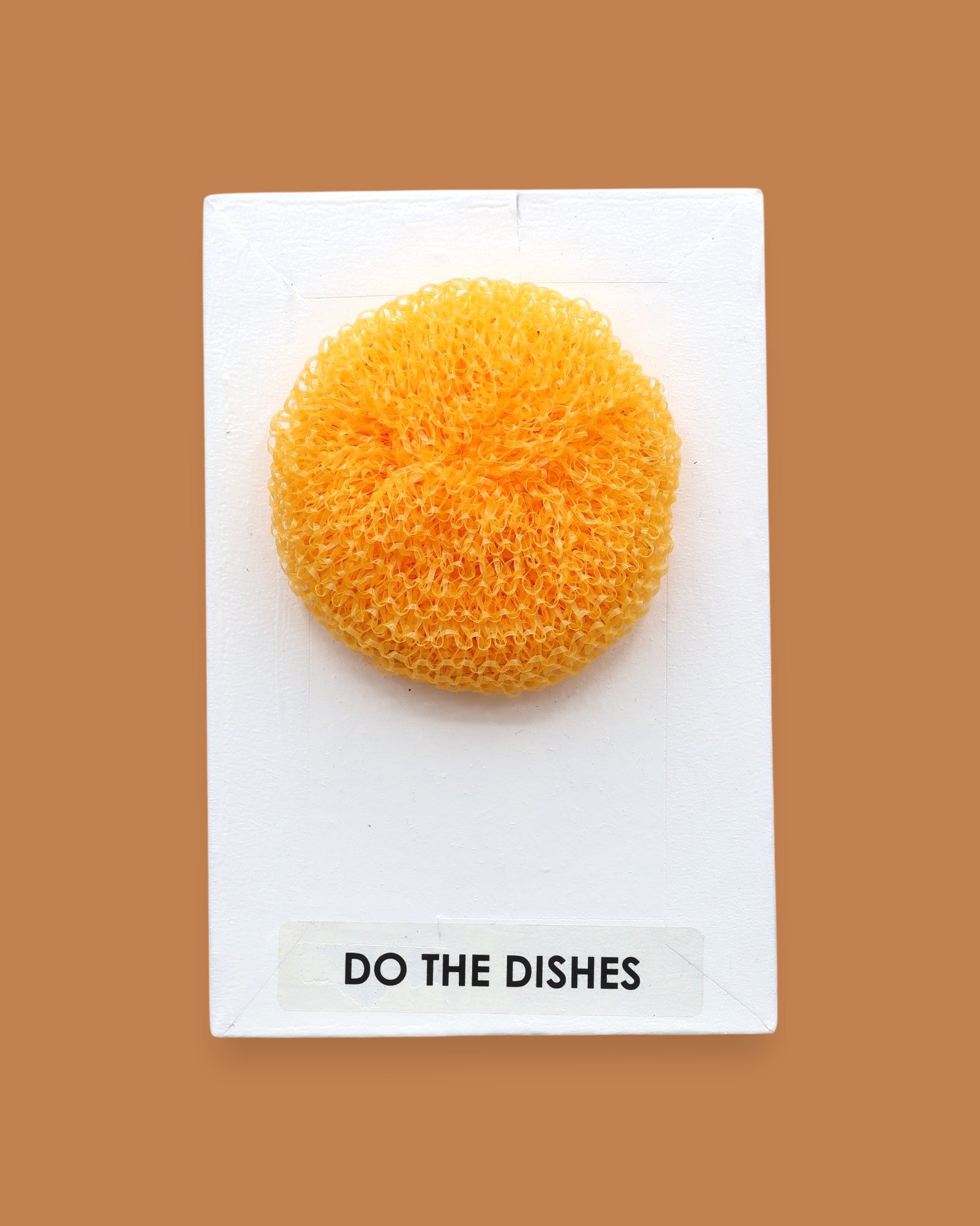The Do The Dishes symbol with a textured cleaning sponge embedded horizontally. Labeled "Do The Dishes" in black font on white backing at the bottom of the symbol. The background of the symbol is white. 