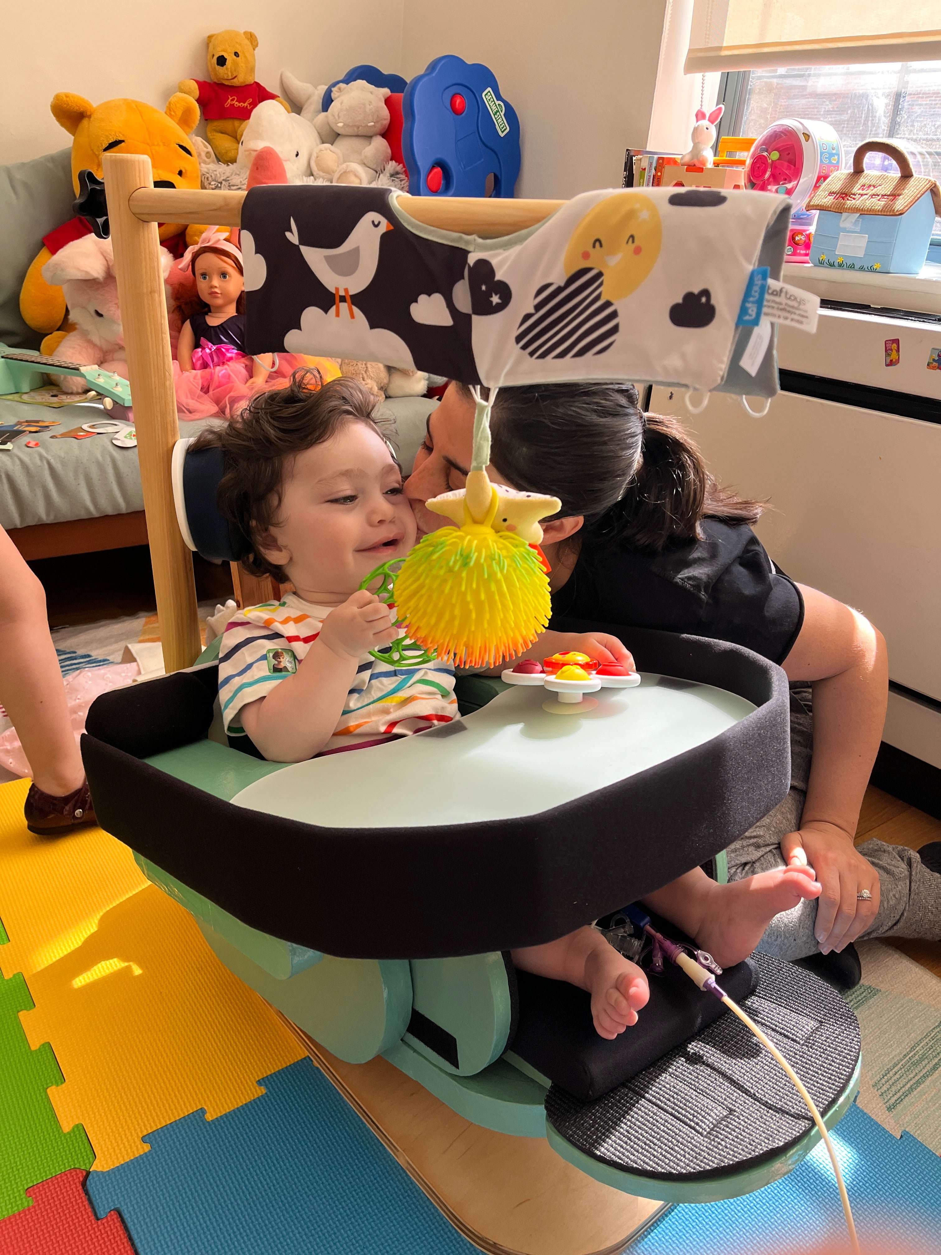 A young child sitting in a floor sitter, made by Adaptive Design, looking upward with an open mouth as if about to react to something fun. In front of the child is a colorful toy with bright yellow and red parts. His mother is seated close by planting a kiss on his cheek. The setting is a children’s playroom  In the background, there are shelves filled with stuffed animals and toys, giving the room a playful and cheerful feel. 
