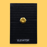 The Elevator high contrast tactile symbol with a flourescent yellow rectangular frame that holds two gold-plated pushable elevator buttons embedded vertically. Labeled "Elevator" in white font on clear backing the bottom of the symbol. The background symbol is black.  