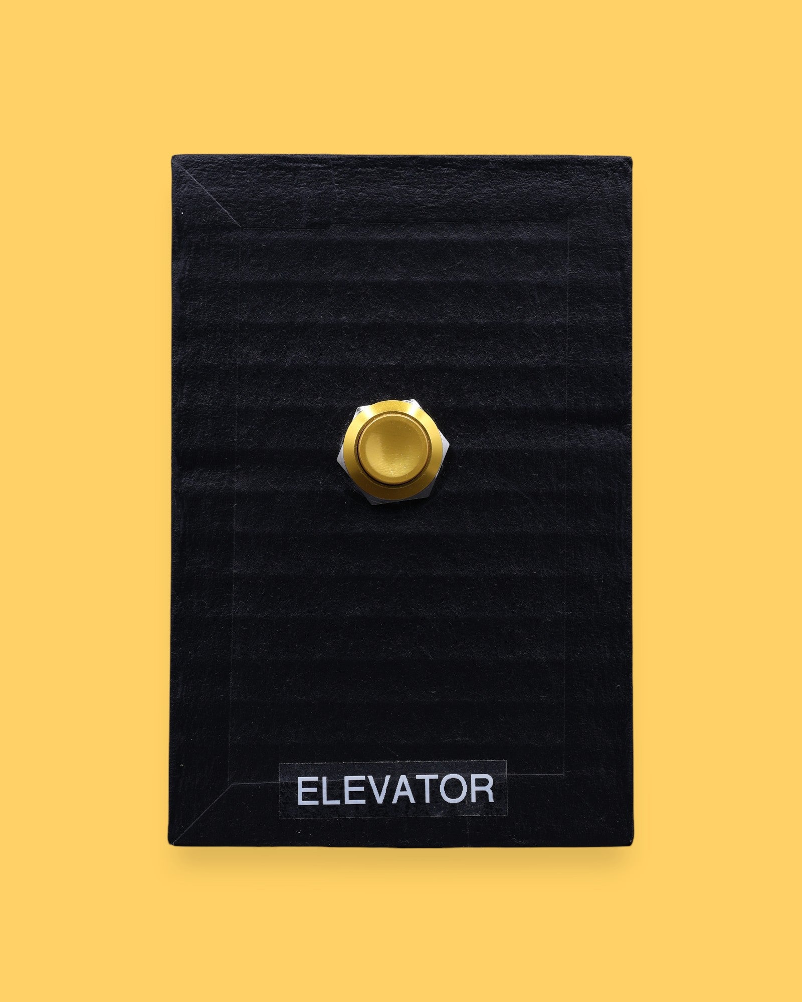 The Elevator high contrast tactile symbol with a flourescent yellow rectangular frame that holds two gold-plated pushable elevator buttons embedded vertically. Labeled "Elevator" in white font on clear backing the bottom of the symbol. The background symbol is black.  