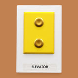 The Elevator symbol with a flourescent yellow rectangular frame that holds two gold-plated pushable elevator buttons embedded vertically. Labeled "Elevator" in black font on white backing at the bottom of the symbol. The background symbol is white.  