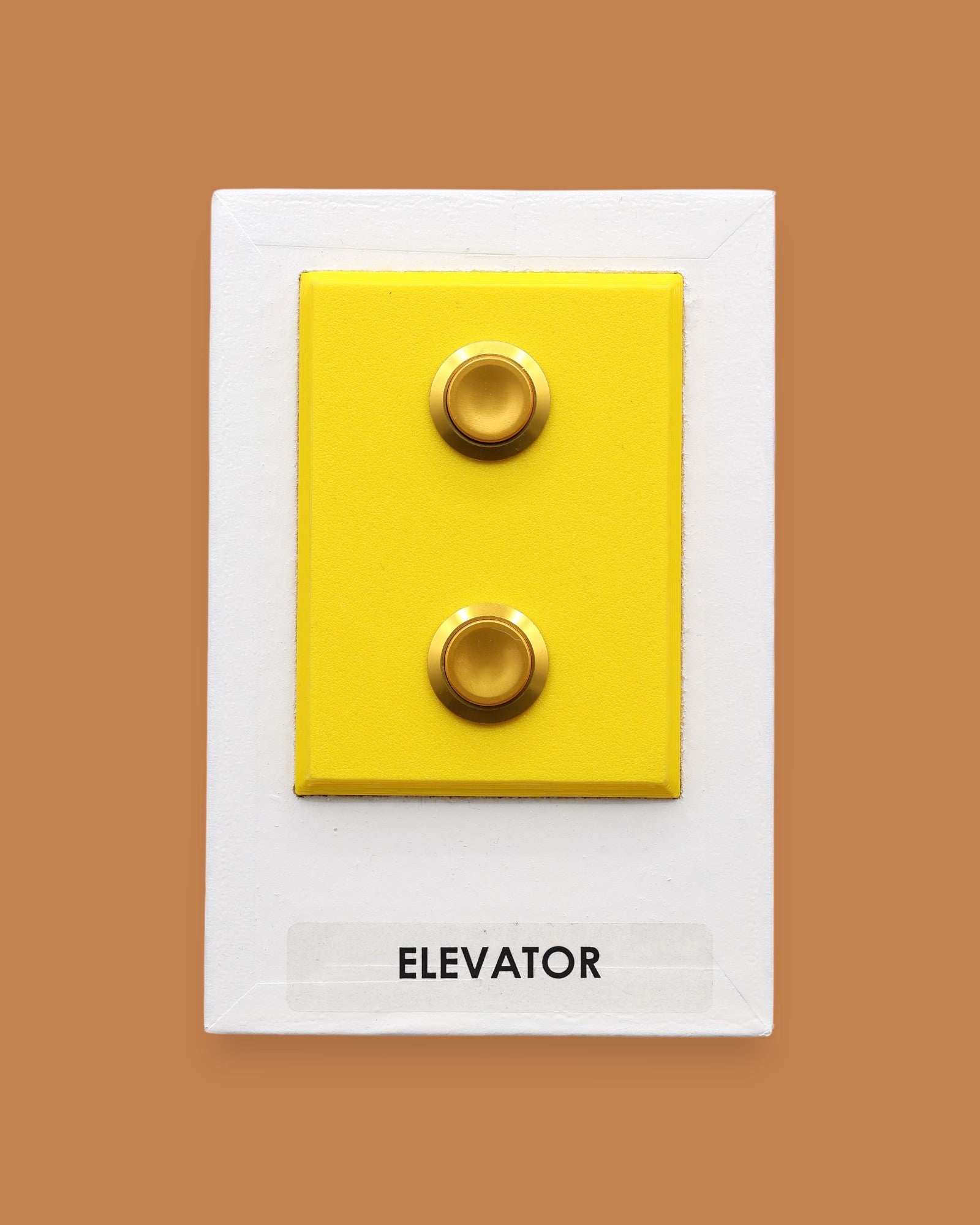 The Elevator symbol with a flourescent yellow rectangular frame that holds two gold-plated pushable elevator buttons embedded vertically. Labeled "Elevator" in black font on white backing at the bottom of the symbol. The background symbol is white.  