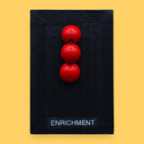 <p>The Enrichment tactile symbol consists of three stacked red beads embedded vertically. Labeled "Enrichment" in white font on clear backing at the bottom of the symbol. The background of the symbol is black.