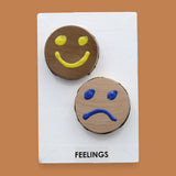 The Feelings symbol with raised handpainted happy and sad face circles embedded diagonally. Labeled "Feelings" in black font on white backing. The background of the symbol is white. 
