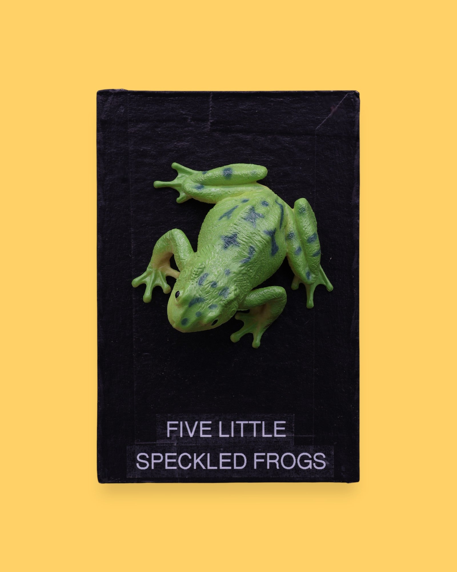 The Five Little Speckled Frogs symbol is a 3" frog toy figure embedded in the center of the cue on top of a recessed wooden base. These plastic frogs come in a variety of designs and colors. The background of the card is black.
