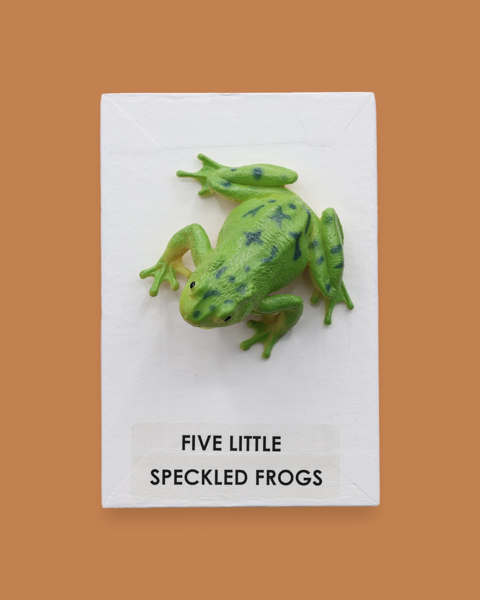 The Five Little Speckled Frogs symbol is a 3" frog toy figure embedded in the center of the cue on top of a recessed wooden base. These plastic frogs come in a variety of designs and colors. The background of the card is white.