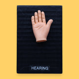 The Hearing-hand high contrast tactile symbol has embedded in the center a small hand shape on a black background. Labeled "Hearing-hand" in white font and clear backing.