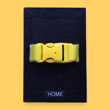 The Home high contrast tactile symbol with a bright yellow closed buckle strap. Labeled "Home" in white font at the bottom of symbol on clear backing. The background of the symbol is black.