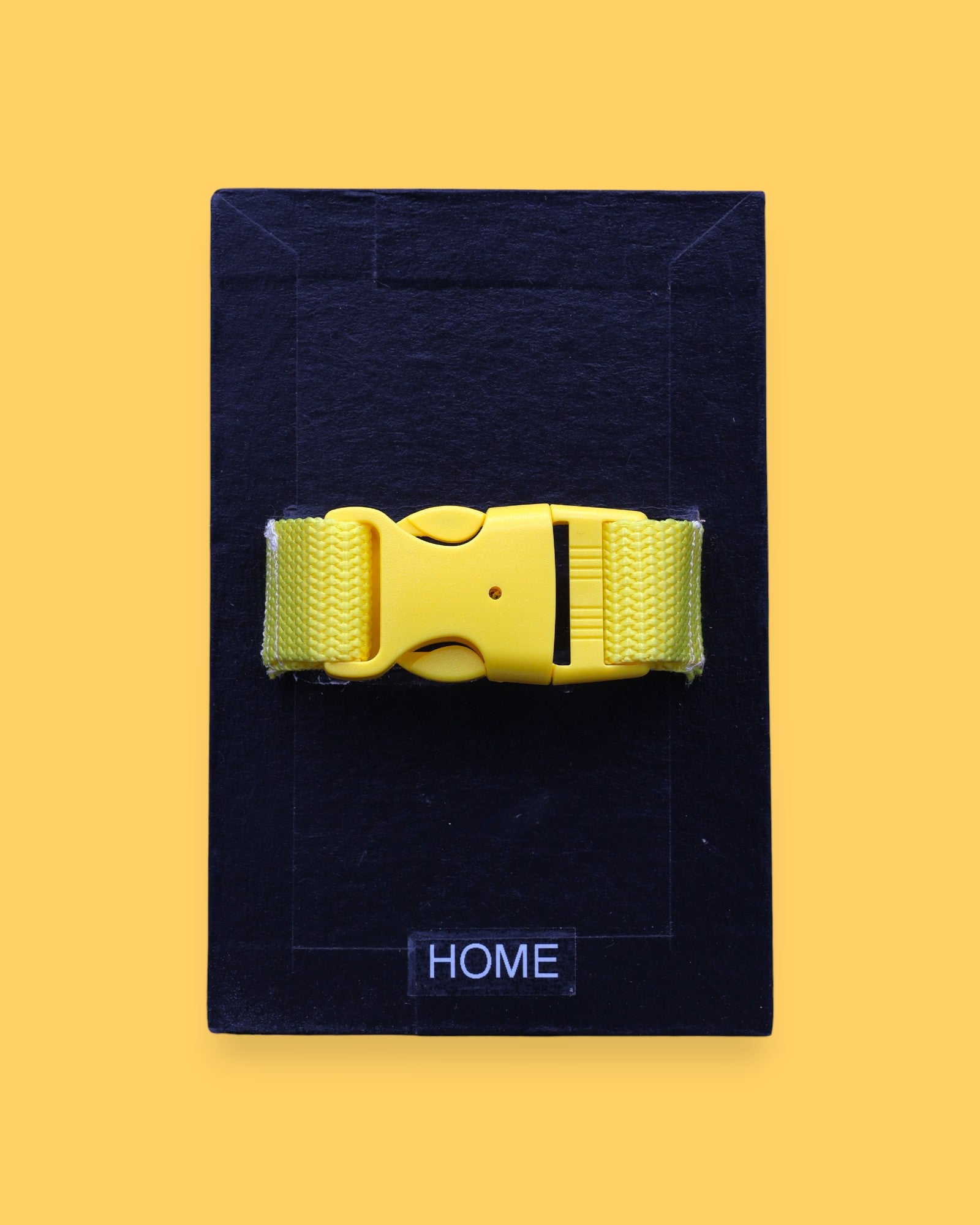 The Home high contrast tactile symbol with a bright yellow closed buckle strap. Labeled "Home" in white font at the bottom of symbol on clear backing. The background of the symbol is black.