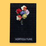 The Horticulture symbol consists of three dried small colorful flowers embedded as a bouquet. Labeled "Horticulture" in white font on clear backing at the bottom of symbol. The background of the symbol is black. 
