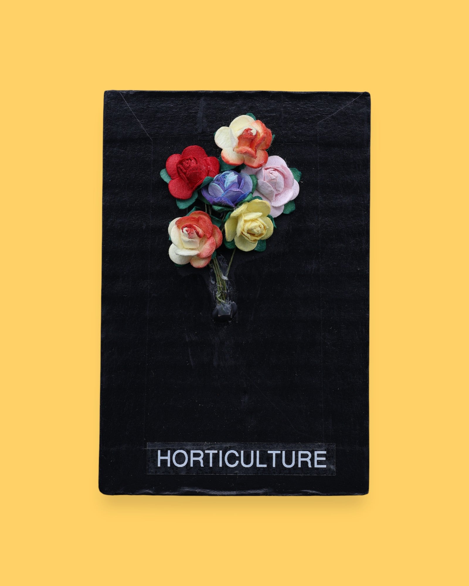 The Horticulture symbol consists of three dried small colorful flowers embedded as a bouquet. Labeled "Horticulture" in white font on clear backing at the bottom of symbol. The background of the symbol is black. 