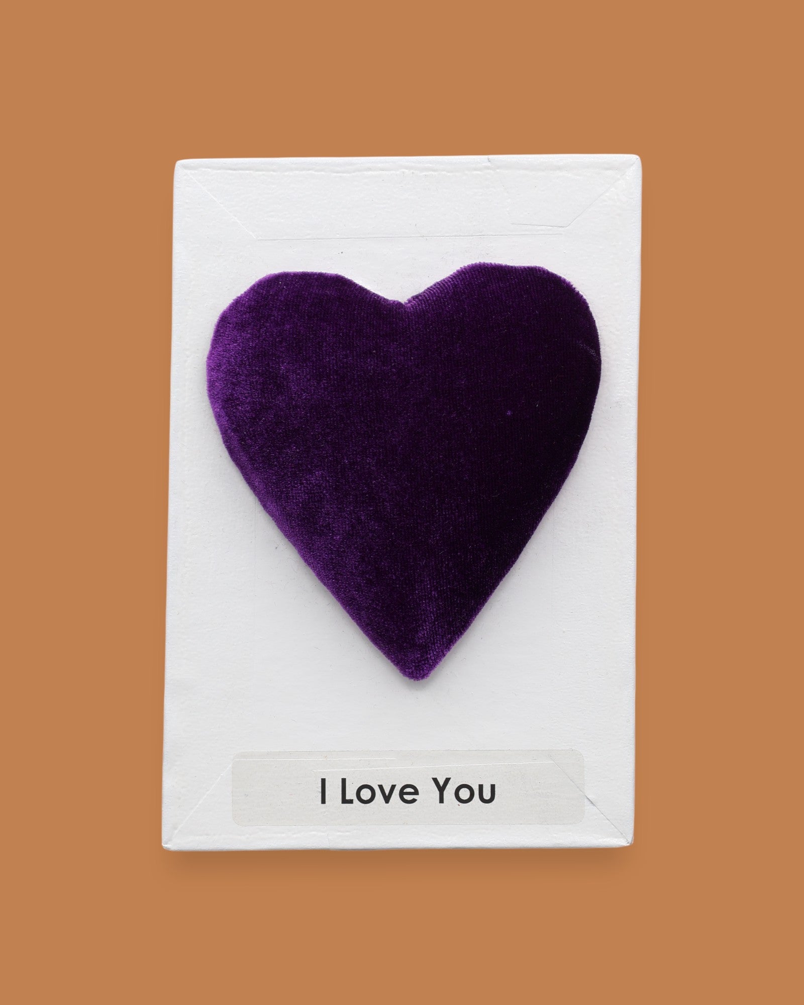 The I Love You symbol with a purple velvet heart attached in the center, labeled "I Love You" in black font at the bottom of symbol on white backing. The background of the symbol is white.