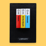 The Library high contrast tactile symbol with three mini book spines in red, yellow and blue. Labeled "Library" in white font at the bottom of symbol on clear backing. The background of the symbol is black. 
