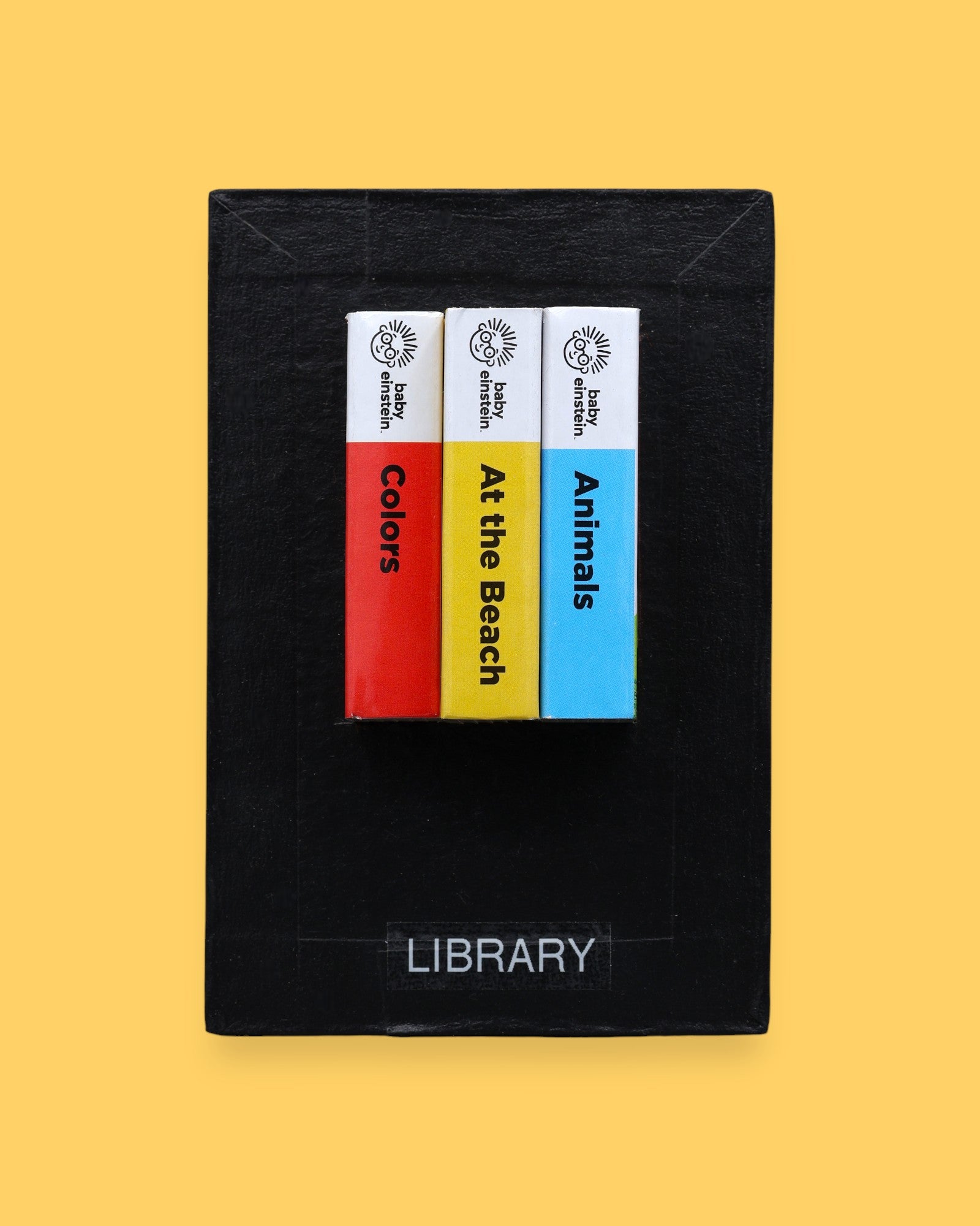 The Library high contrast tactile symbol with three mini book spines in red, yellow and blue. Labeled "Library" in white font at the bottom of symbol on clear backing. The background of the symbol is black. 