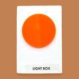The Light Box Plastic Shape symbol with a smooth circular piece embedded in the center. Labeled "Light Box" in black font on white backing at the bottom of the symbol. The background of the symbol is white. 
