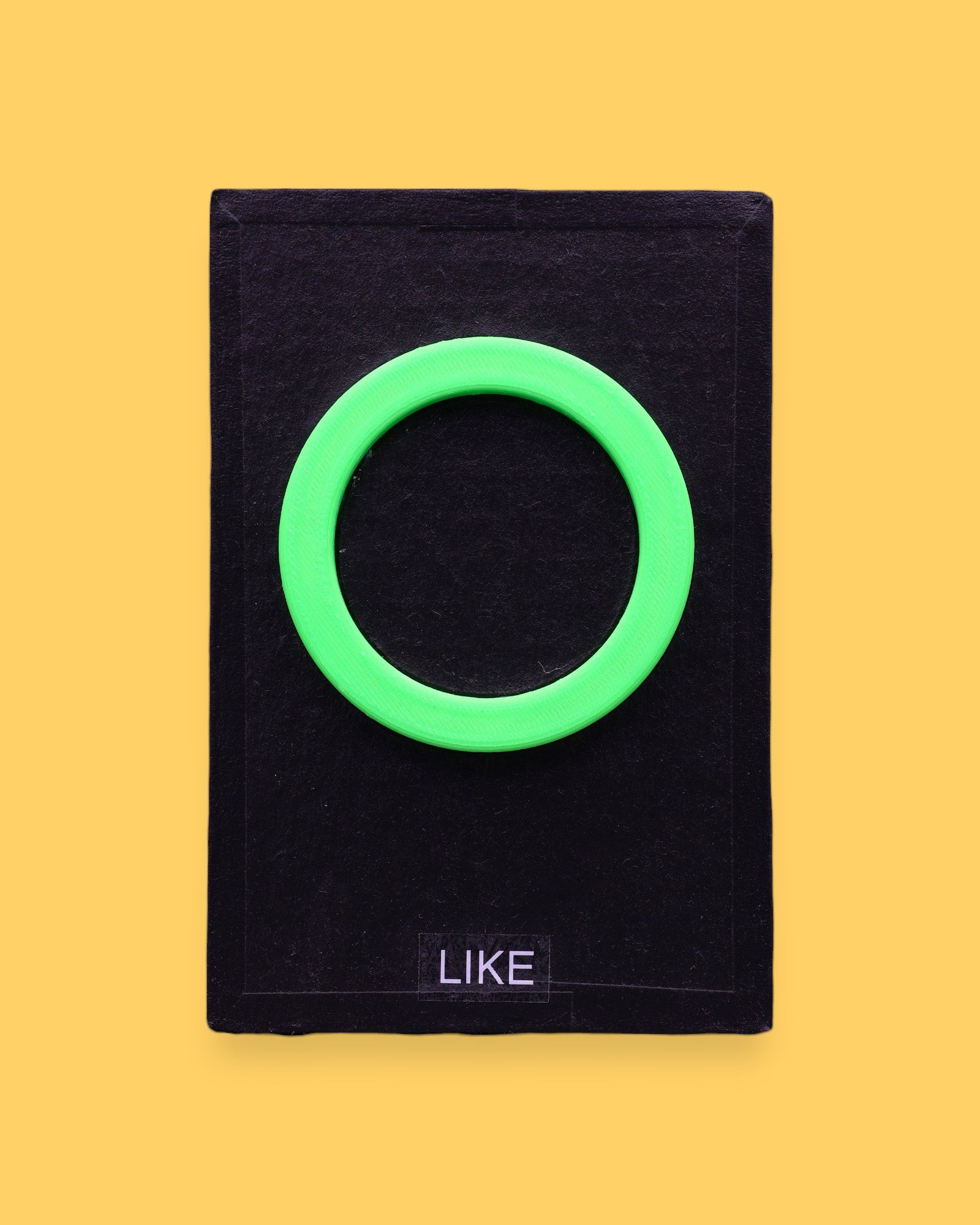 The Like high contrast tactile symbol card for Like with a neon 3D printed PLA filament green ring embedded in the center. Labeled "Like" in white font at the bottom of symbol on clear backing. The background of the symbol is black. 